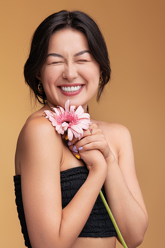 Cheerful young Asian female smiling with closed eyes and touching shoulder with fresh gerbera flower during spa session against brown background