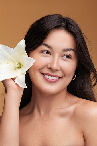 Glad Asian woman with bare shoulders looking away with smile and putting white lily flower into black hair during spa session against brown background