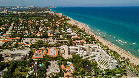 Aerial 4K Drone View of Beach-Front Housing Overlooking a Vibrant Beach Filled with Blue Ocean Waves, Rocks, Seaweed, Beach Umbrellas, Surf Boards, & People on the Sandy Shoreline in Palm Beach, Florida at Midday During Springbreak 2023