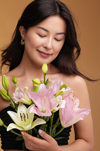 Glad Asian woman with bare shoulders admiring bunch of fresh lily flowers while standing against brown brown background