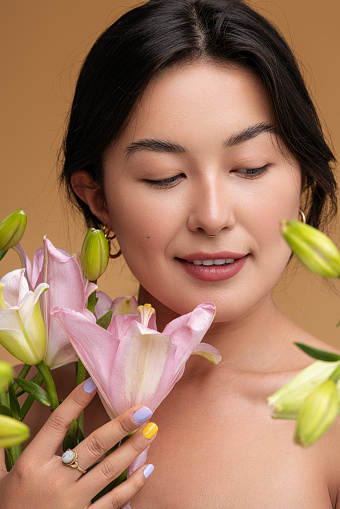 Optimistic young Asian female smiling and touching fresh lily flowers with delicate petals against brown background