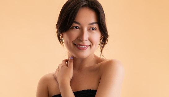 Optimistic young Asian female with bare shoulders touching neck and looking away with smile during spa session against beige background