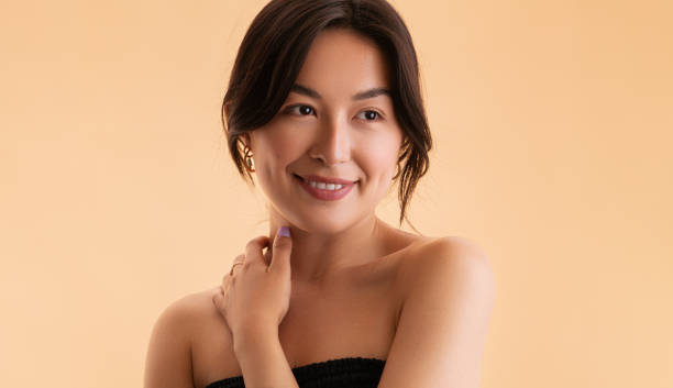 Glad Asian woman touching neck Optimistic young Asian female with bare shoulders touching neck and looking away with smile during spa session against beige background strapless stock pictures, royalty-free photos & images