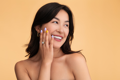 Cheerful young Asian female with bare shoulders looking away with smile and touching cheek during skin care routine against beige background