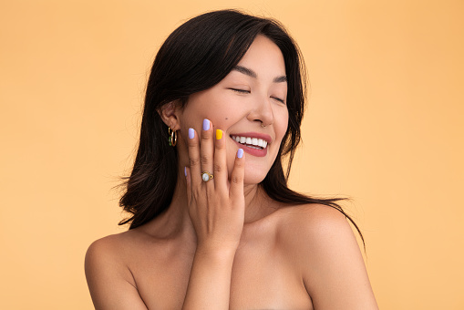 Optimistic young Asian female with bare shoulders touching clean cheek and smiling with closed eyes during skin care routine against beige background