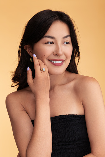 Cheerful Asian female with bare shoulders looking away with smile and touching clean cheek during skin care routine against beige background