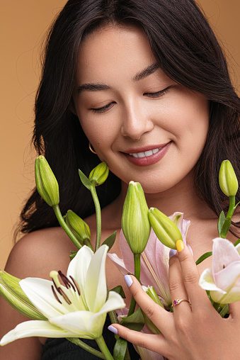 Happy young Asian female smiling and touching buds of fresh lily flowers against brown background