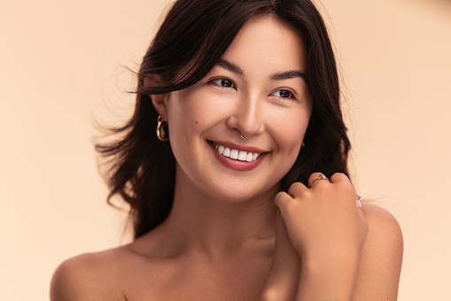 Happy young Asian female touching bare shoulder with clasped hands and looking away with smile against beige background