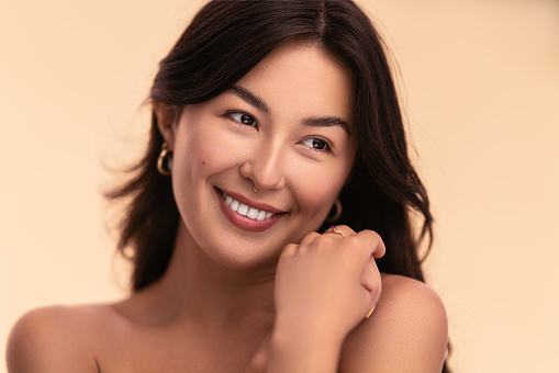 Young Asian female with clean face and gorgeous hair smiling while looking away on beige background