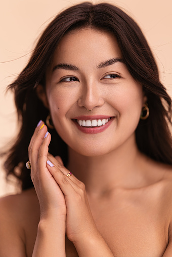 Happy young Asian female with dark hair clasping hands and looking away with smile during spa session against beige background