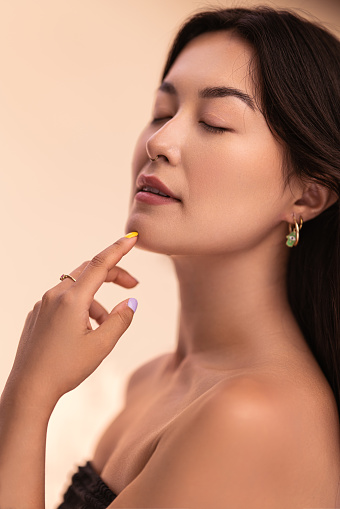 Tranquil young Asian female with bare shoulders closing eyes and touching chin during skin care routine against beige background