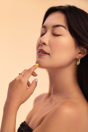 Young Asian woman with bare shoulders closing eyes and touching chin during skin care routine against beige background
