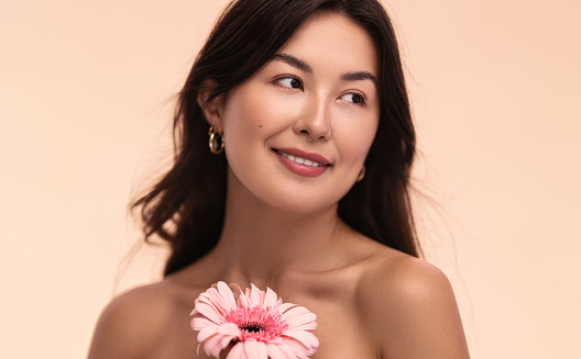 Young Asian female with fresh gerbera flower smiling and looking away during skin care routine against beige background