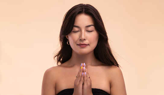 Calm young Asian female clasping hands and closing eyes while meditating against beige background