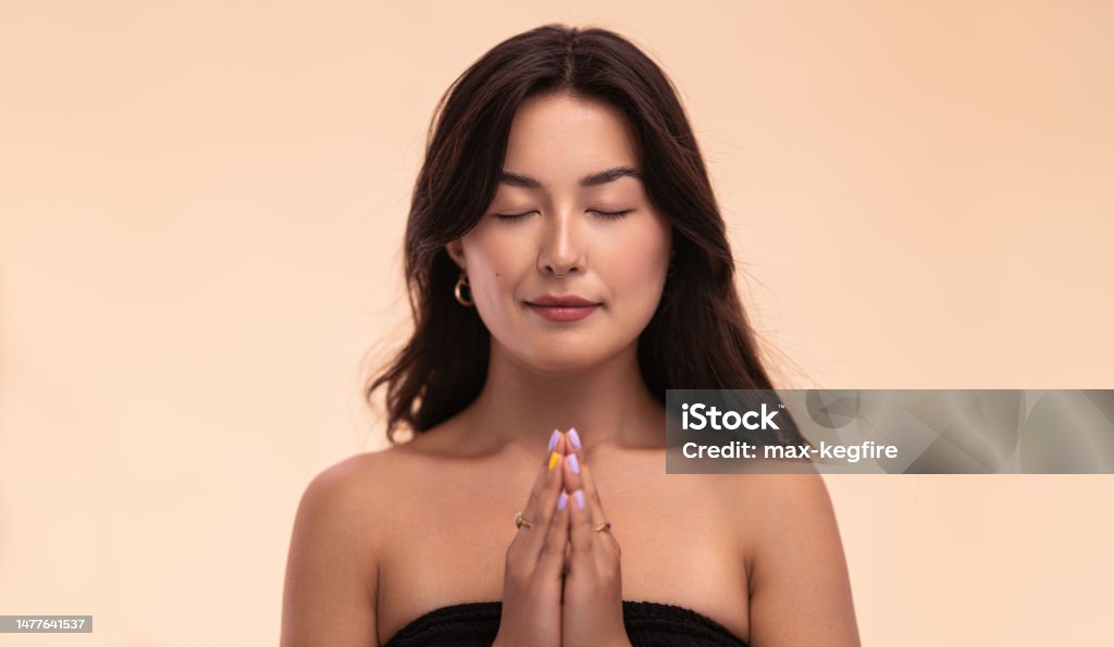 Asian woman meditating with clasped hands Calm young Asian female clasping hands and closing eyes while meditating against beige background Mindfulness Stock Photo
