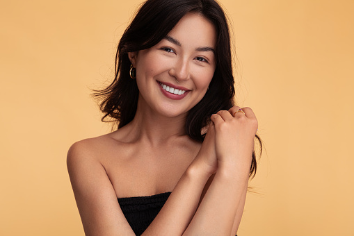 Optimistic young Asian woman with bare shoulders clasping hands and looking at camera with smile against beige background