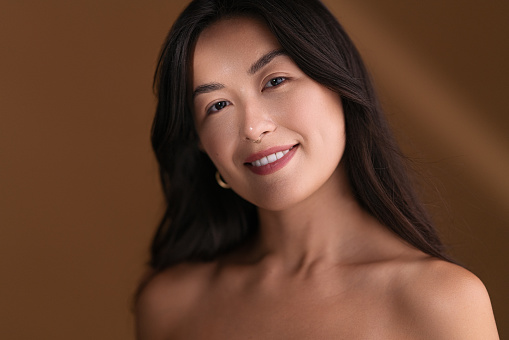 Happy young Asian female with bare shoulders smiling and looking at camera while standing in sunbeam against brown background