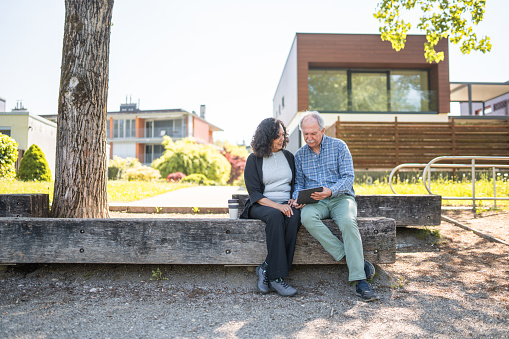 Aged, mixed couple sitting on a  wooden bench, looking at something on a digital tablet. Both dressed sportively. Full length shot, residential area  in the background. Looking away.