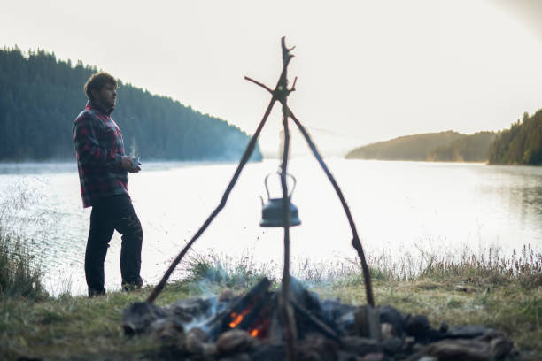 Young man standing by the campfire enjoying the beauty of nature on a camping vacation in the morning.