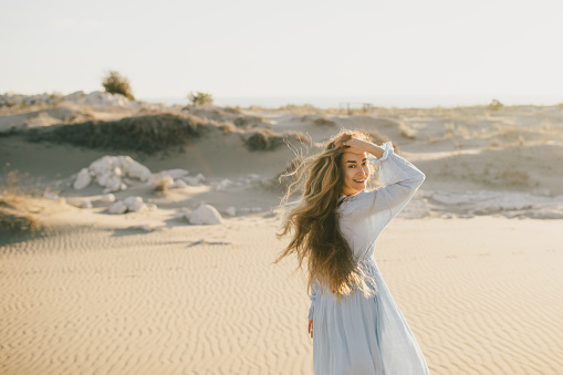 A beautiful woman with long hair in a stylish blue dress poses barefoot in the desert sands.