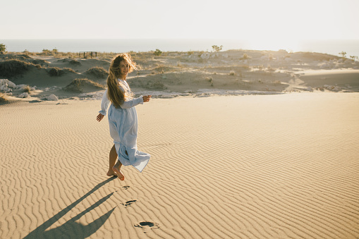 A beautiful woman with long hair in a stylish blue dress poses barefoot in the desert sands.