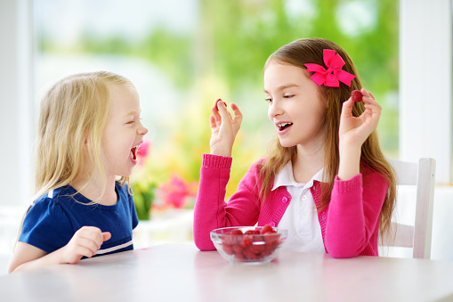 Two pretty little girls eating raspberries at home. Cute children enjoying their healthy fresh organic fruits and berries. Kids having fun together.