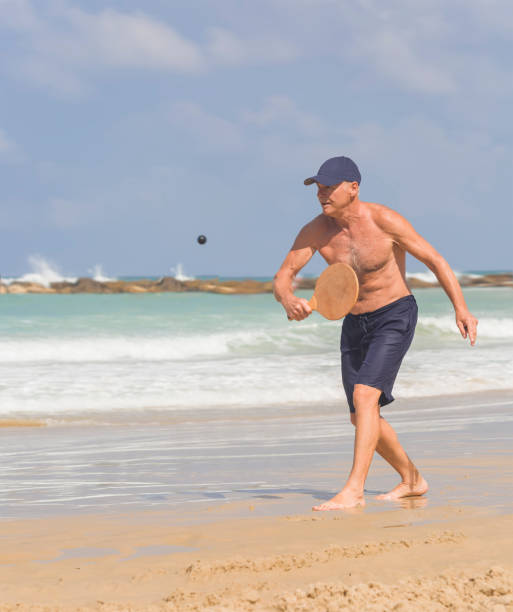 ハンサムな成熟した男がビーチでマトコットをしながらボールを打つ - matkot ストックフォトと画像