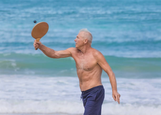 un bell'uomo maturo colpisce una palla mentre gioca a matkot sulla spiaggia - matkot foto e immagini stock