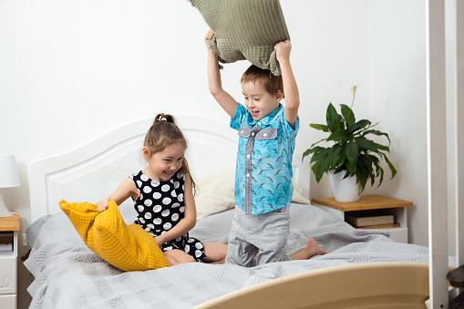 Pillow fight. Mischievous preschooler children jumping on bed and hitting each other with pillows. Boy and girl play together. Active games for siblings at home
