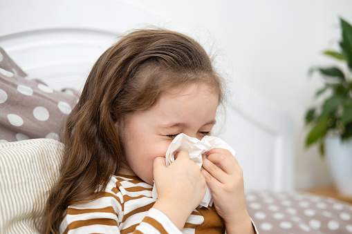 Little Caucasian girl with influenza is sitting in bed and sneezes, blowing her nose. The child is sick, runny nose