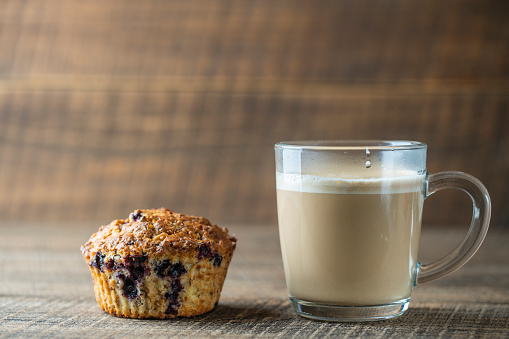 Delicious muffin with blueberries and cappuccino glass cup on a wooden table, close up. Fresh cupcake and coffee for breakfast, copy space