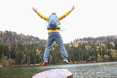 Happy male traveler with backpack jumping near forest lake while reaching goal back view closeup