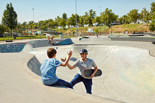 Smiling man wearing helmet and holding skateboard high-fiving boy sitting on edge of bowl as they enjoy weekend recreation together.