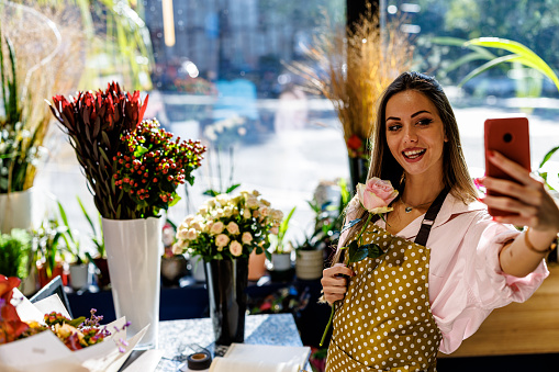 A beautiful female florist is taking a self-portrait photo with a mobile phone in a modern flower store.