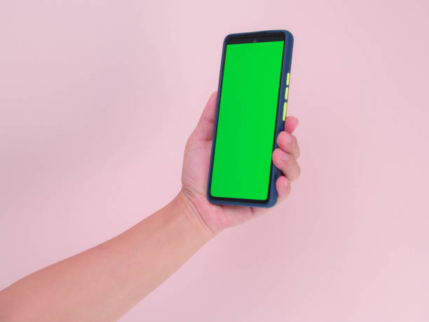 istock Human hand holding mobile smartphone with green screen in vertical position isolated on pink background. clipping path 1477716636