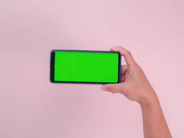 istock Human hand holding mobile smartphone with green screen in horizontal position isolated on pink background. clipping path 1477716652