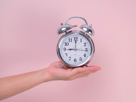 Female hand holding classic alarm clock on pink background. Close up of alarm clock in hand.