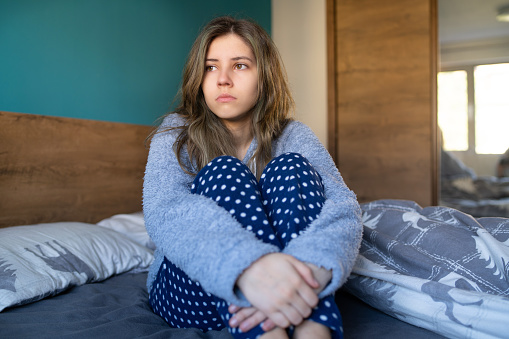A young woman is sitting on her bed with her hands wrappred around her legs looking sad and lonely, wearing her pyjamas