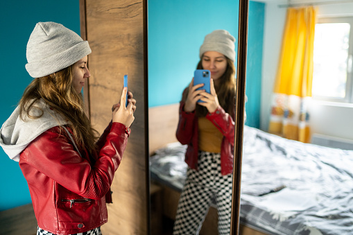 A young woman is holding her mobile phone up to take a photo of herself in front of a large mirror in a modern bedroom