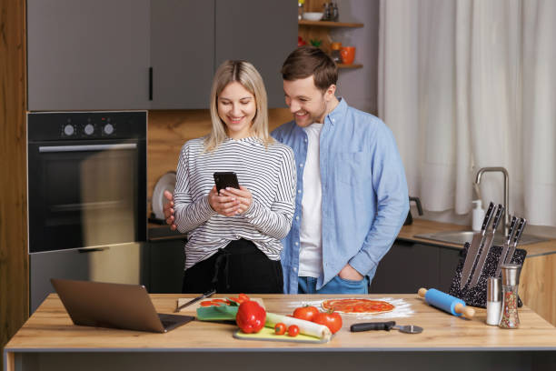 istock Smiling loving couple cooking salad together while standing on a kitchen at home and using mobile phone. Cute couple man and woman taking photo of food and taking pictures together while cooking. 1477721922