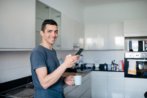 Portrait of smiling man having coffee and using phone in the kitchen at home.