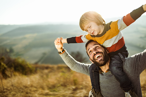 Happy single father having fun while carrying his small boy on shoulders during autumn day on a field. Copy space.