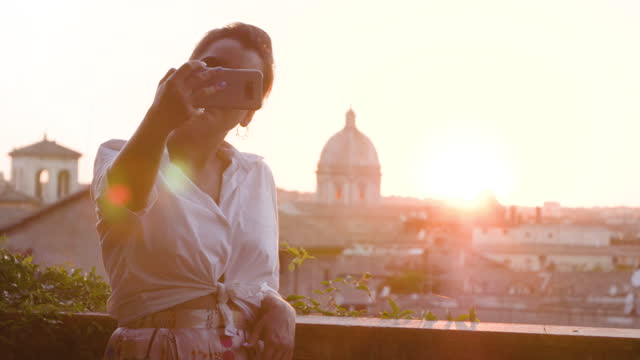 Young woman talking to camera in front of Rome cityscape from high balcony viewpoint in campidoglio. Landmarks and domes panoramic view at sunset.