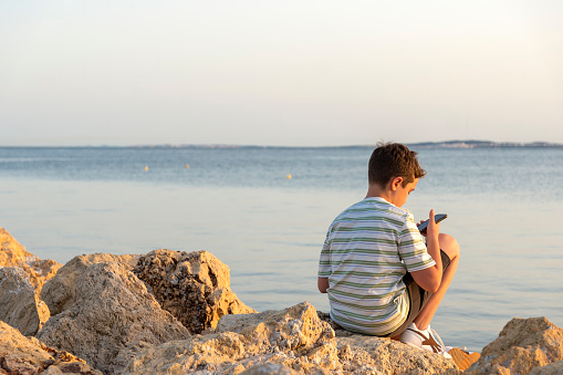 A teenager uses his smartphone while sitting on rocks in front of the sea at sunset.