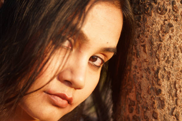 Asian Indian female extreme close up portrait of serious neutral emotion looking into camera in outdoors evening light her head leaning on to a tree stock photo