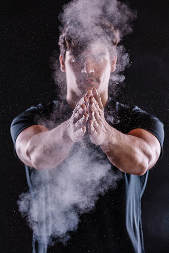 Closeup portrait of young strong sportsman applying chalk powder on hands at studio, isolated over dark black background. Clapping hands with magnesium.