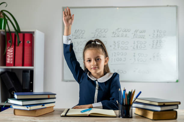 An elementary school student raises her hand because she is ready to answer the teacher's question in the classroom. A smart little girl is sitting at a desk in the classroom. Education concept.