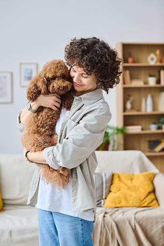 Vertical image of young woman holding dog in her arms and embracing it while standing in the room