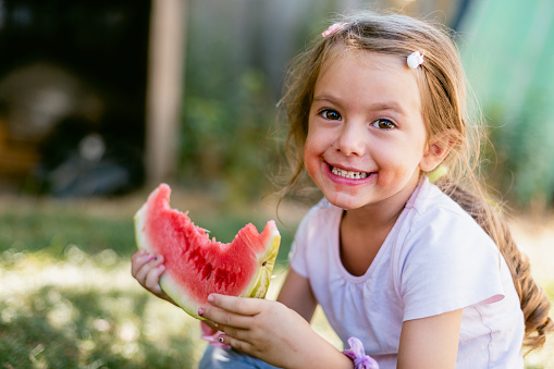 Cute messy little girl eating slice of watermelon on hot summer day