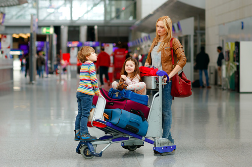 Two little kids, boy and girl, siblings and mother at the airport. Children, family traveling, going on vacation by plane and waiting on trolley with suitcases pushing by woman at terminal for flight